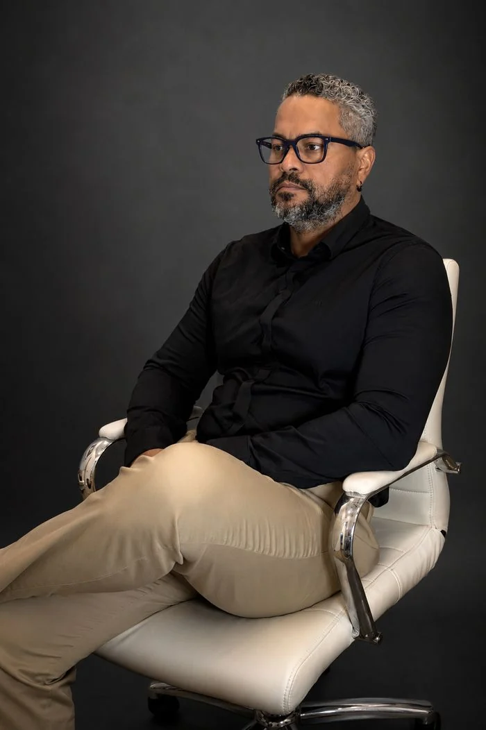 Angel Duran in a black shirt, seated in a white chair, looking at the camera